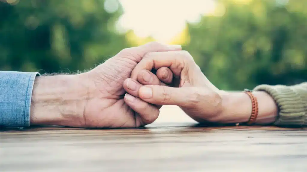Two hands, one male and one female, clasped together on a wooden table, with a blurred green background. The male hand is slightly larger and has a denim cuff, while the female hand is adorned with a bracelet.