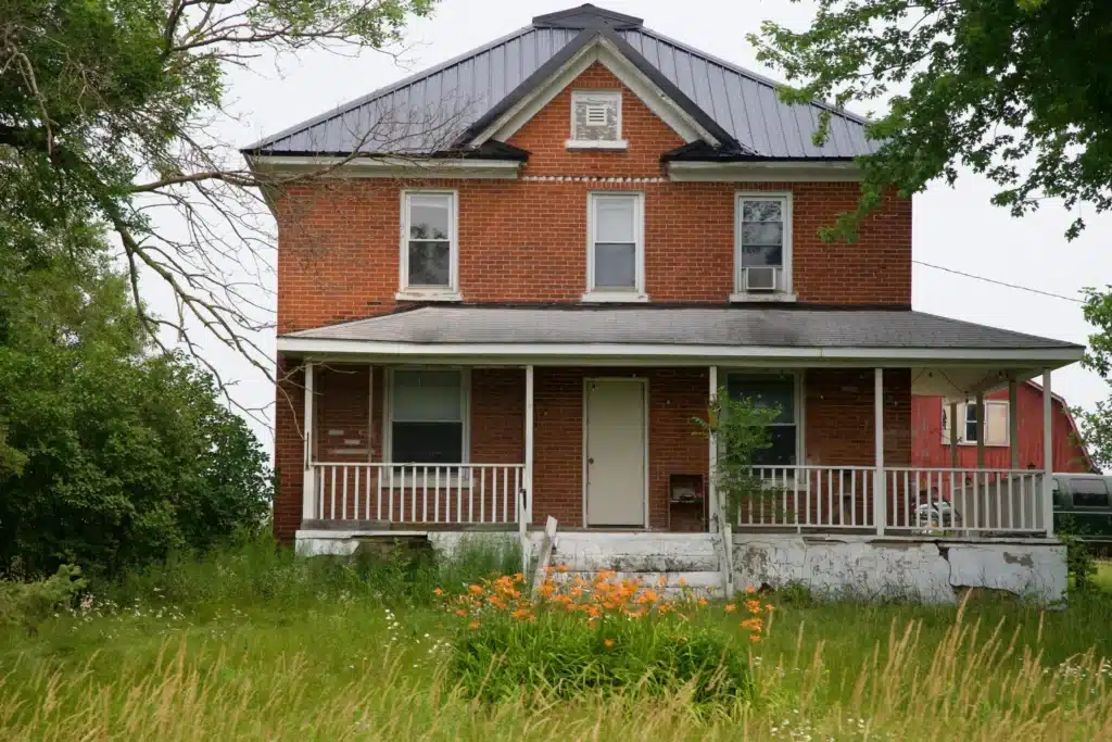 A two-story red brick house with a black roof, featuring a front porch and surrounding greenery. Orange flowers bloom in the foreground, and there is a second structure visible in the background.