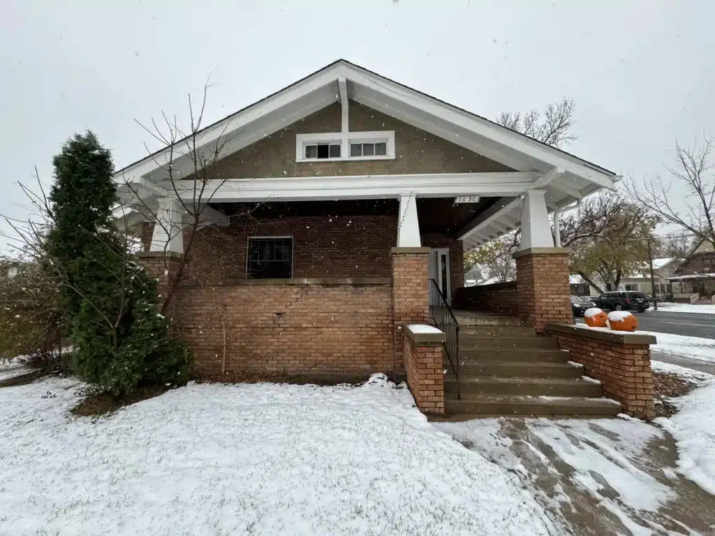 A house with a covered porch, made of brick and wood, surrounded by light snow. There are pumpkin decorations on the steps, and green shrubs on the left side of the image.